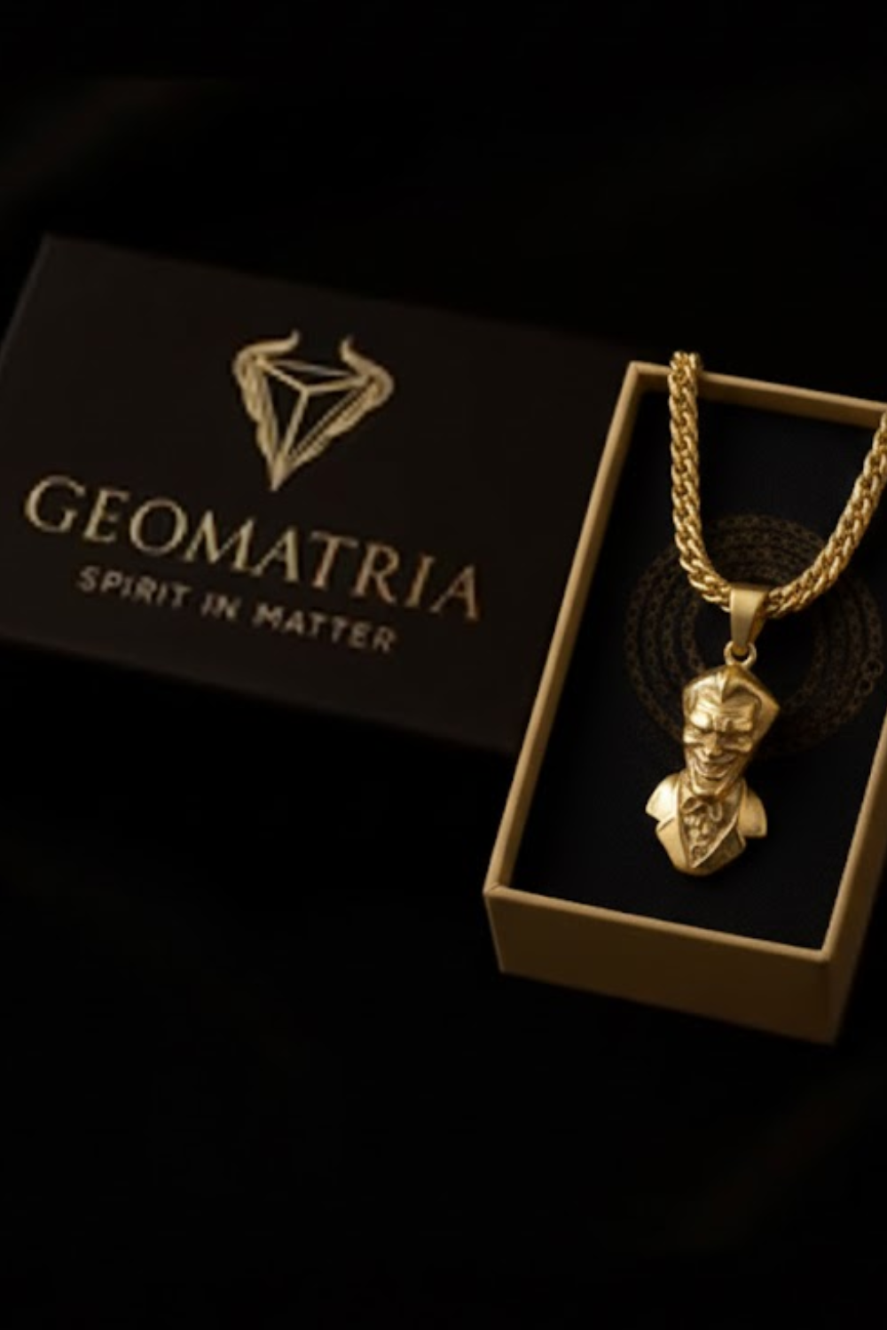 A gold-colored necklace with a pendant is displayed in a black box with the text "Geomatria Spirit in Matter" printed on it.