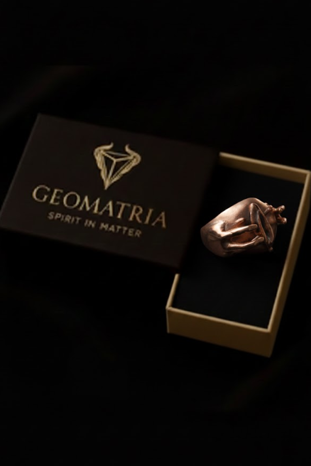 A gold ring is displayed inside a black box with the word "Geomatria" printed on it.