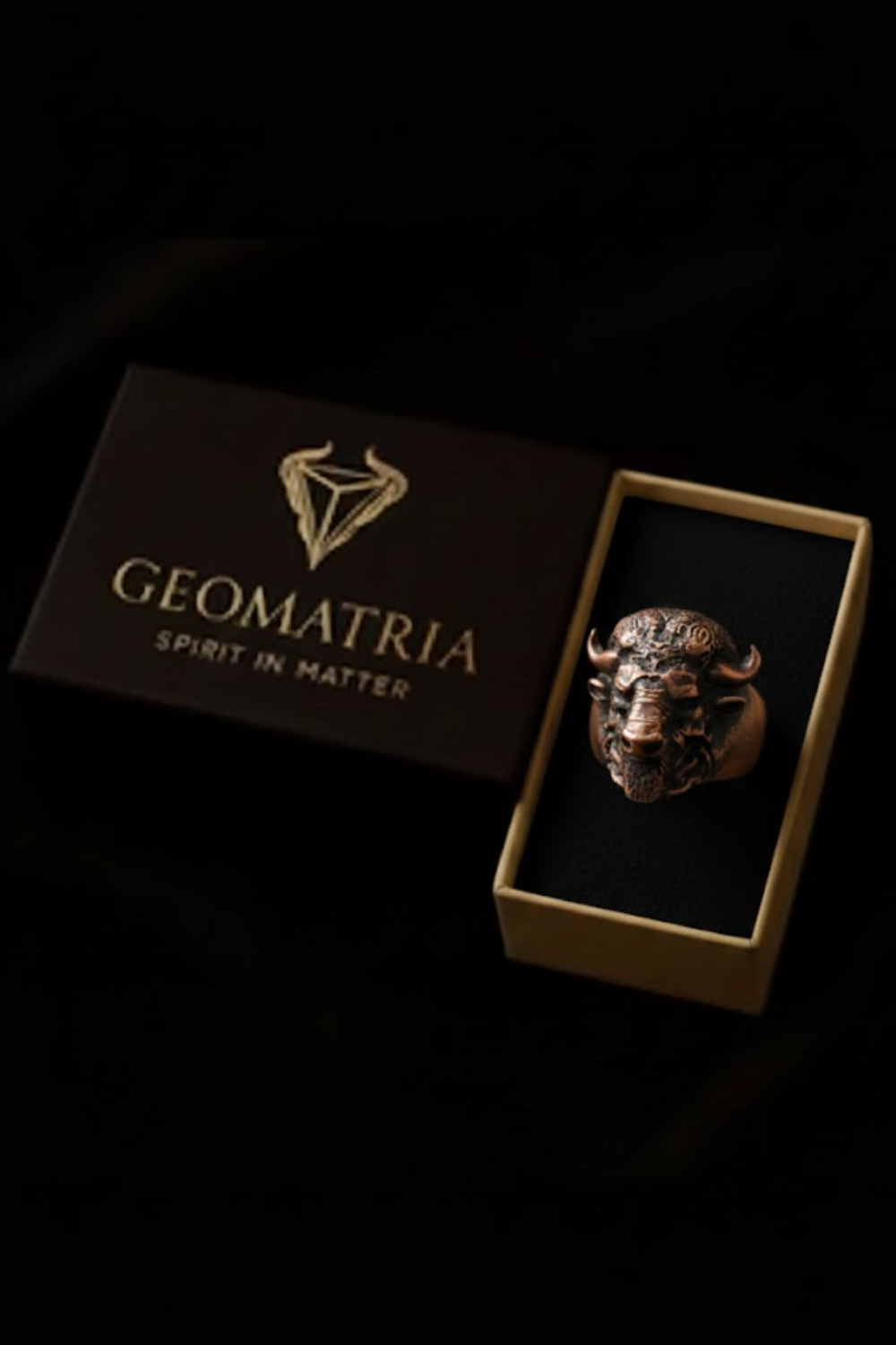 A ring with a bull's head design is displayed in a gold box with the text "Geomatria" and "SPIRIT IN MATTER" printed on it.