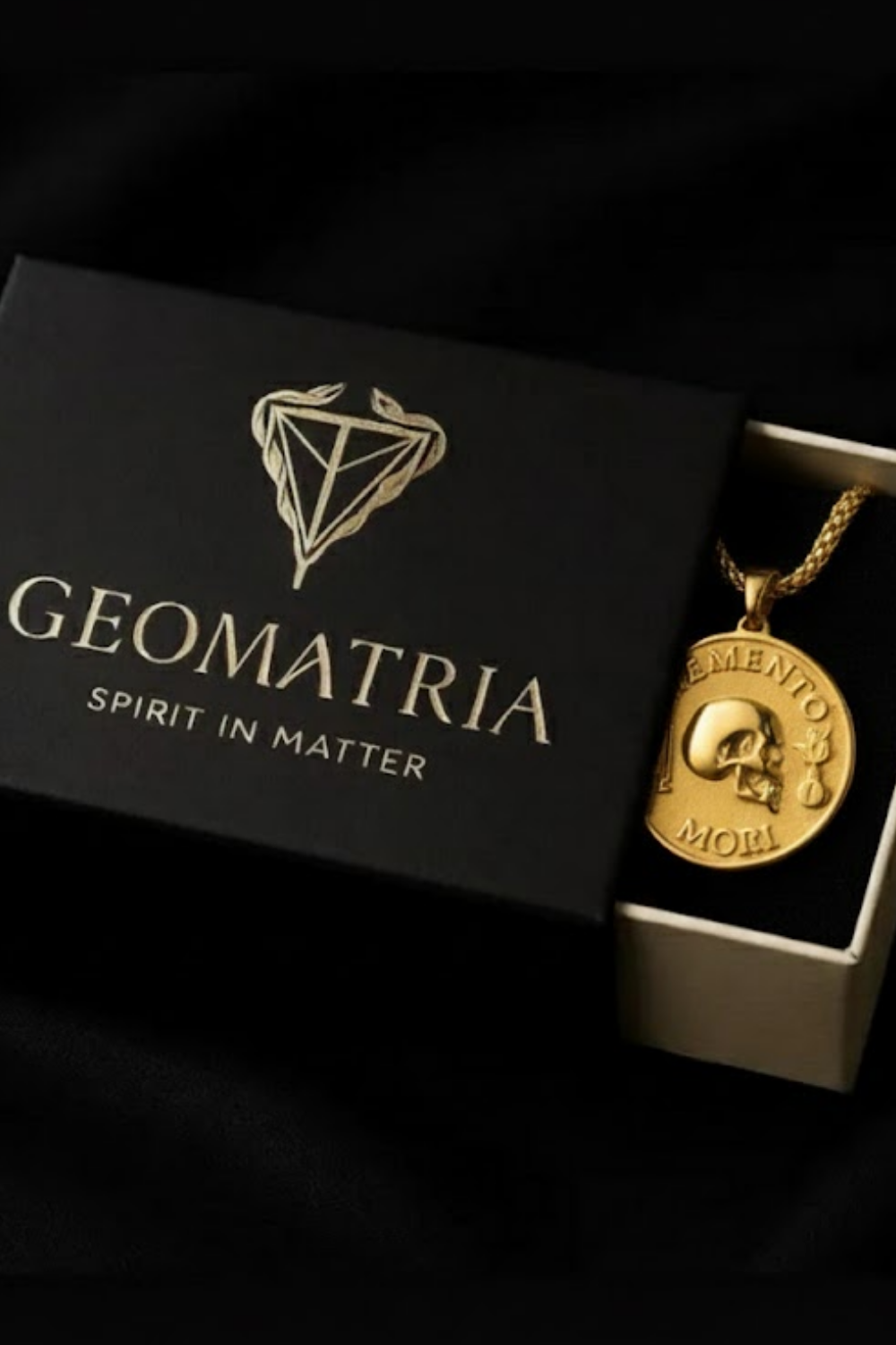 A gold-colored pendant necklace with a skull pendant is displayed in a black box with the text "Geomatria" and "SPIRIT IN MATTERS" printed on it.