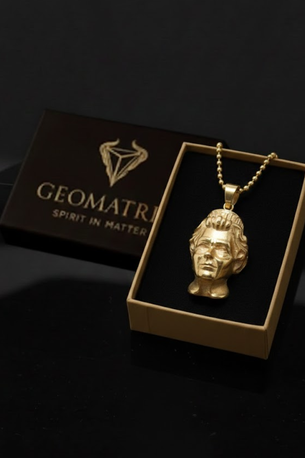 A gold-colored pendant in the shape of a human head is displayed in a gold-colored box with the text "Geometri" and "SPIRIT IN MATTER" printed on it.