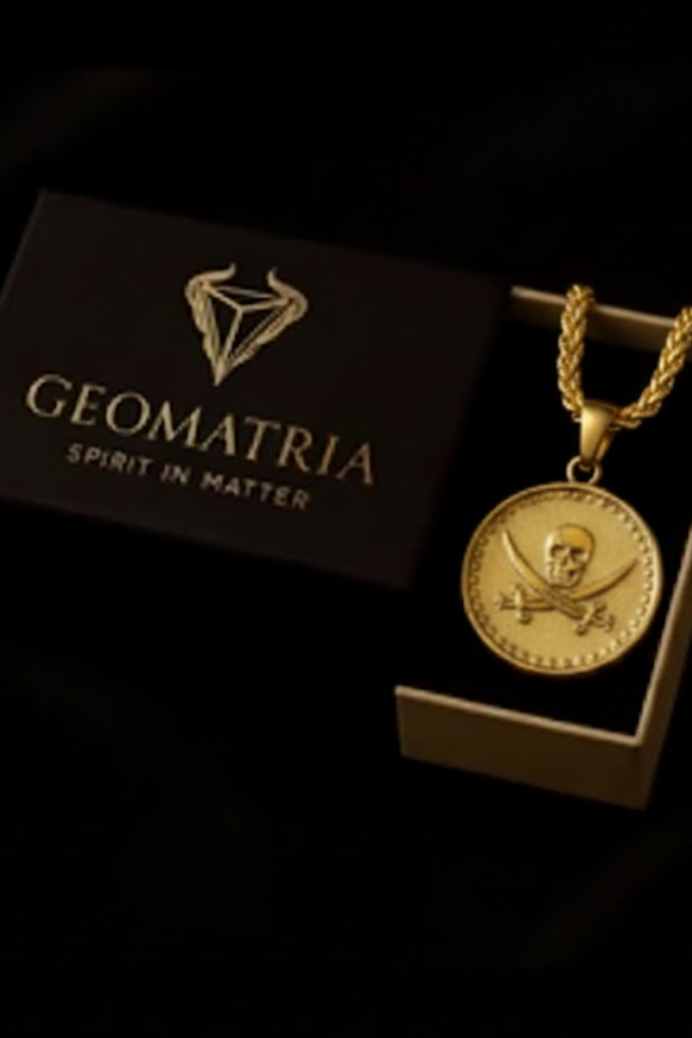 a gold-colored pendant with a skull and crossbones design, displayed in a black box with the text "Geomatria Spirit in Matter" printed on it.