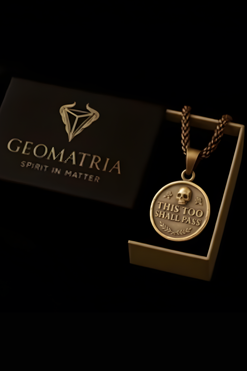 A gold-colored pendant necklace with a skull pendant, displayed in a black box with the Geomatria logo.