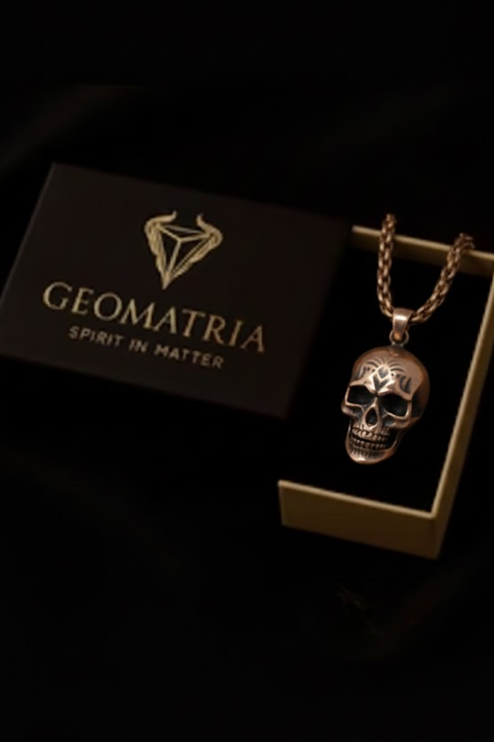 A gold-colored pendant necklace with a skull design is displayed in a black box with the Geomatria logo.
