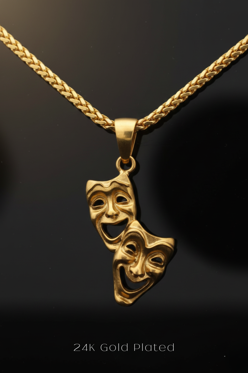 A 24K gold-plated pendant featuring two gold-plated theatrical masks with exaggerated features, hanging from a chain against a dark background.