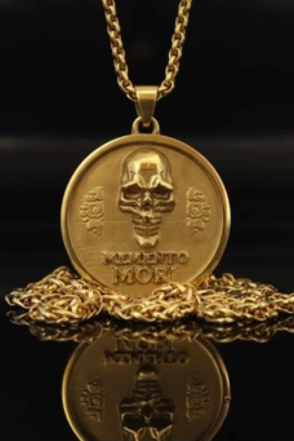 a gold-colored pendant necklace with a circular medallion featuring a skull design and the Latin phrase "Memento mori" (which means "life after death"). The pendant and chain are placed on a reflective surface, creating a visually striking contrast between the gold and black background.