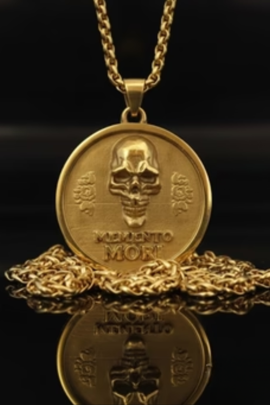 a gold-colored pendant necklace with a circular medallion featuring a skull design and the Latin phrase "Memento mori" (which means "life after death"). The pendant and chain are placed on a reflective surface, creating a visually striking contrast between the gold and black background.