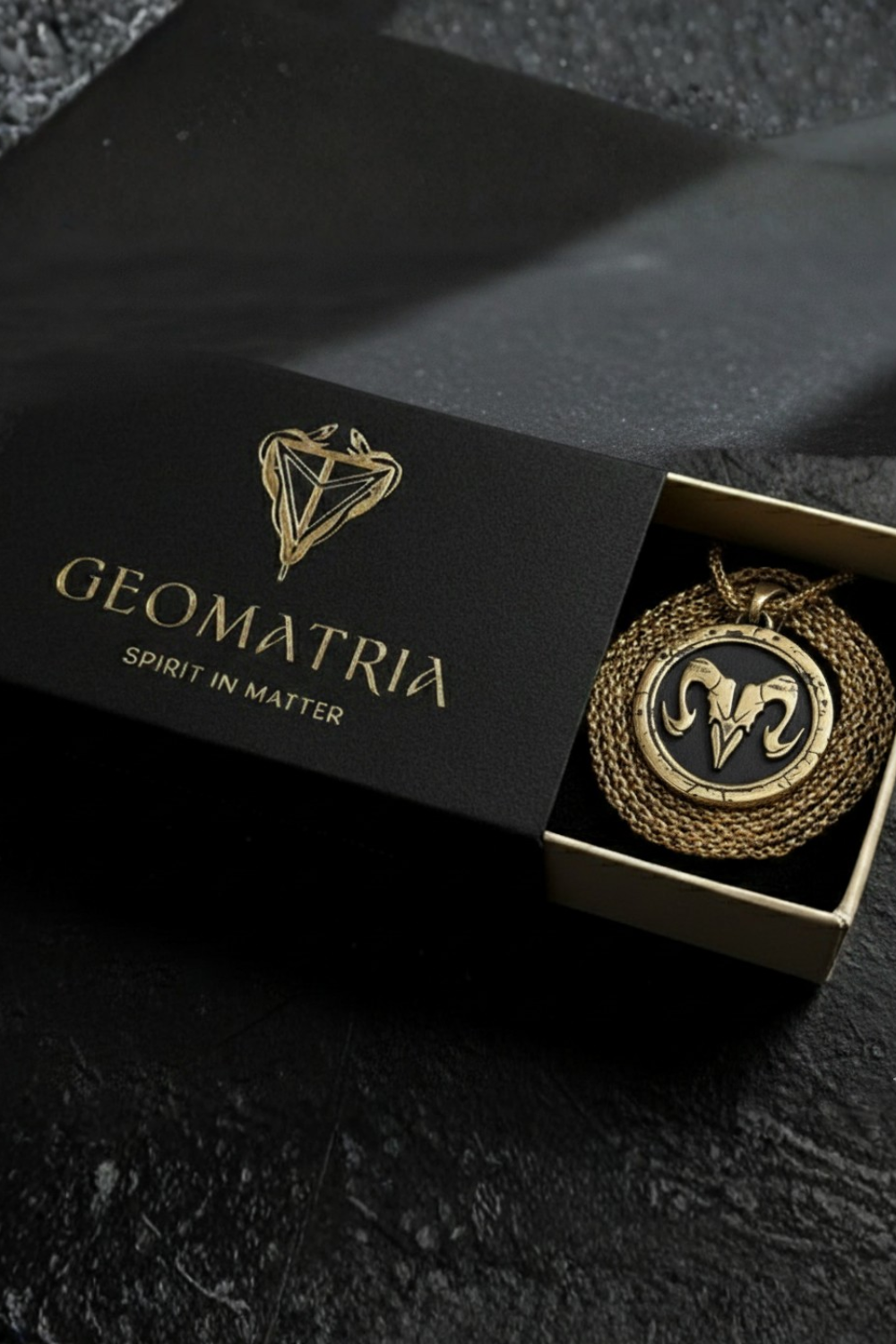 A black box with a gold geometric design and the text "Geomatria" on it, along with a gold chain necklace with a pendant featuring a geometric design.