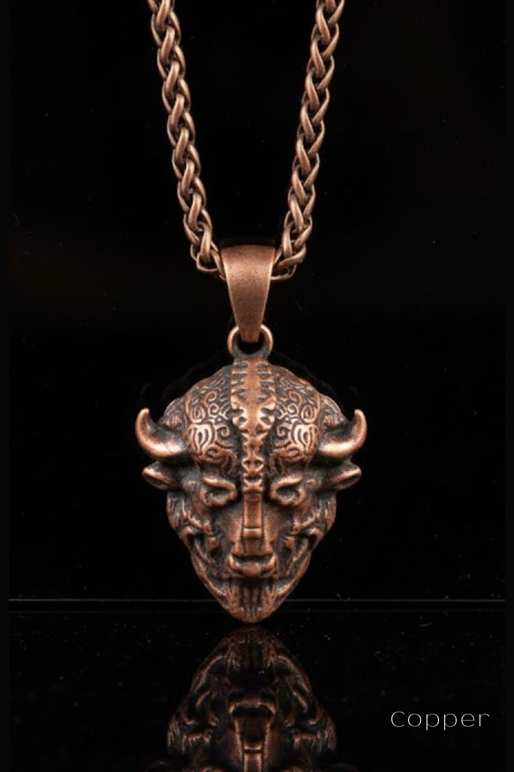 A copper-colored pendant necklace with a detailed, intricate design featuring a stylized, abstract face or mask-like figure.
