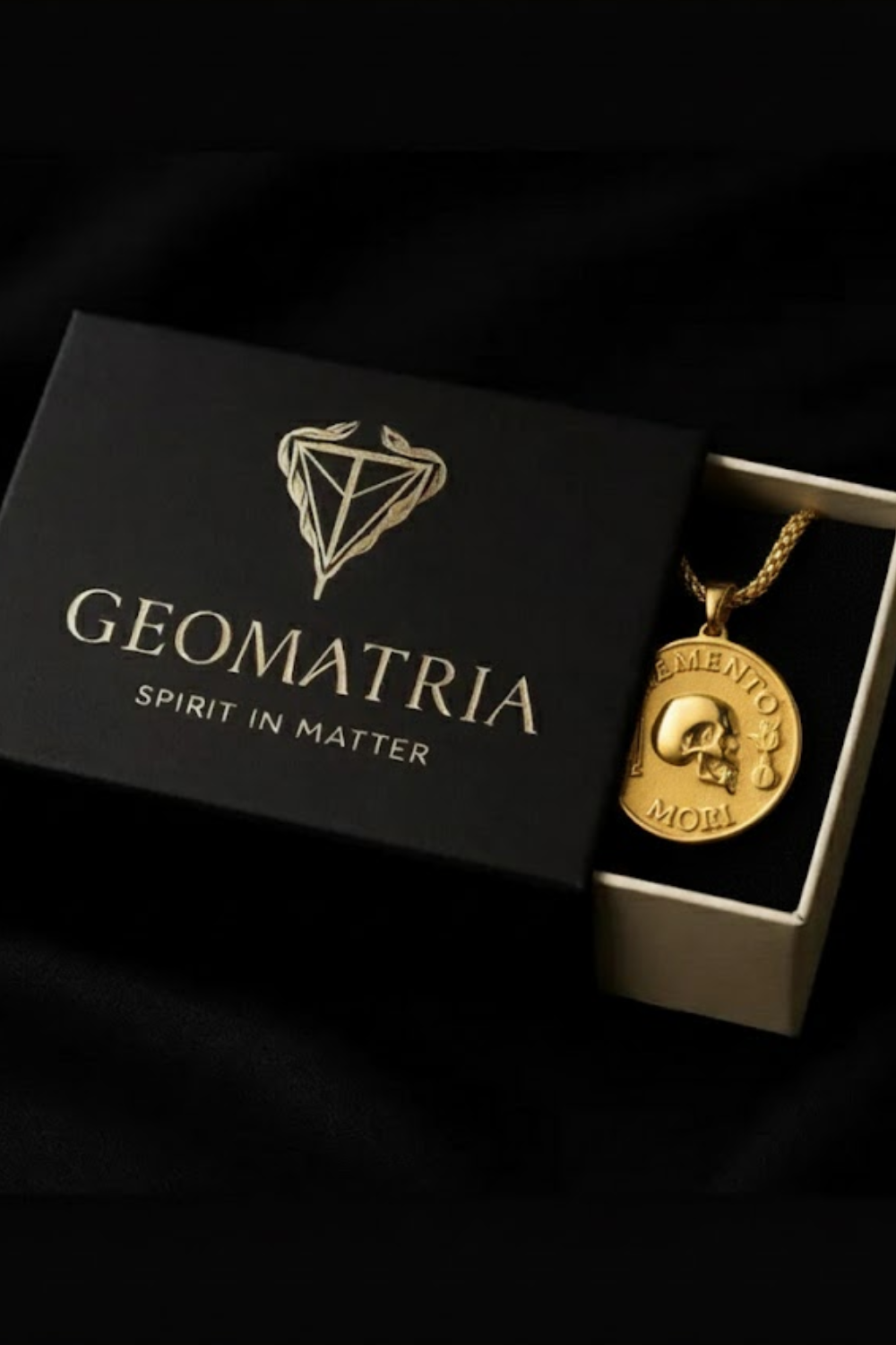 A gold-colored pendant necklace with a skull pendant is displayed in a black box with the text "Geomatria" and "SPIRIT IN MATTERS" printed on it.