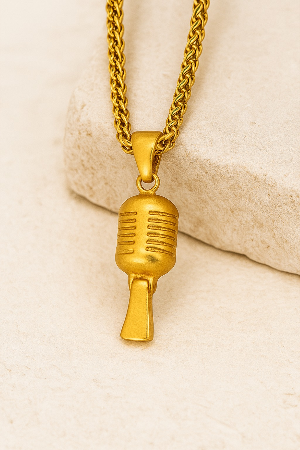 A gold-colored microphone pendant is displayed on a chain necklace against a light-colored background.