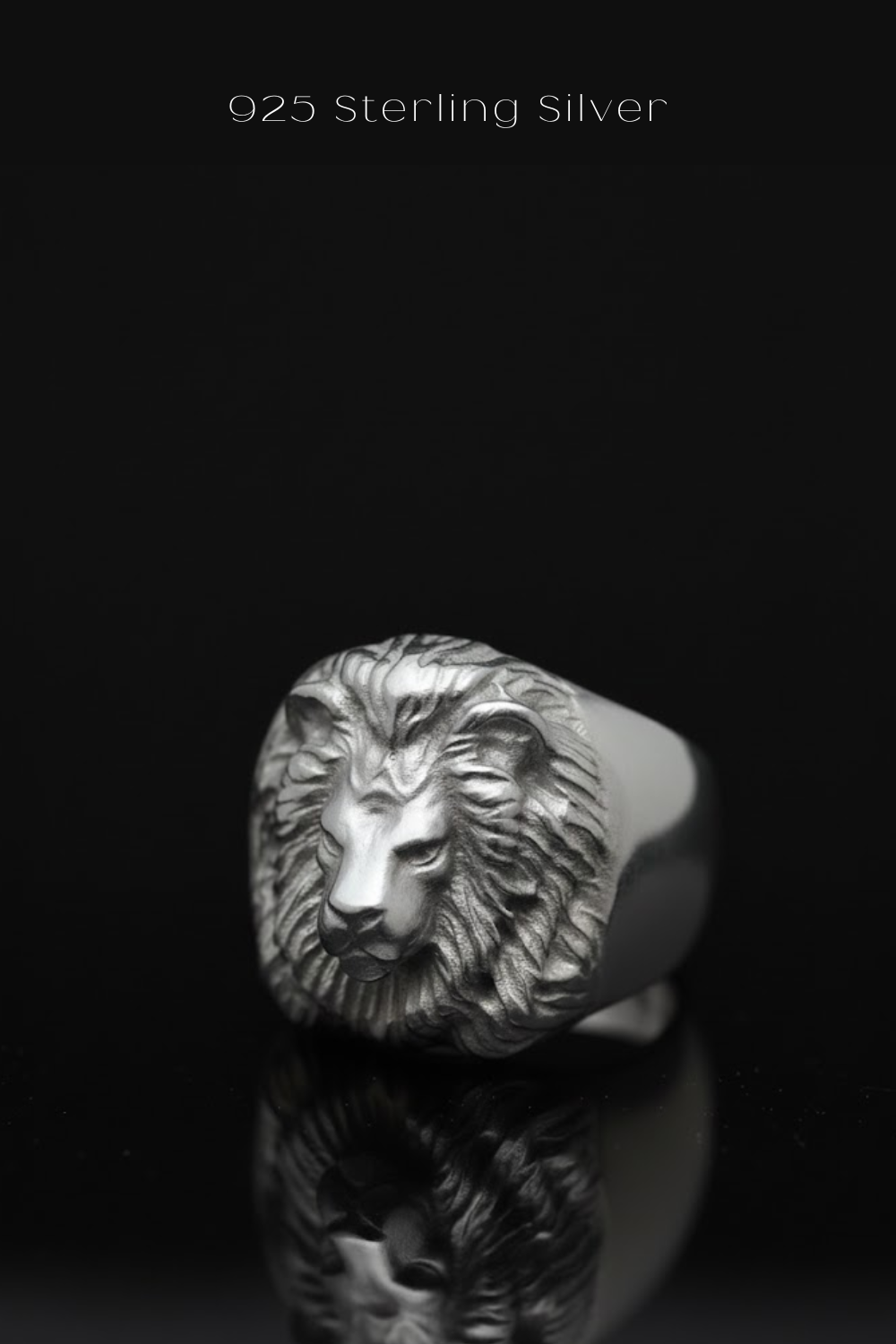 A silver lion head ring is displayed against a black background, with its reflection visible on the surface below.