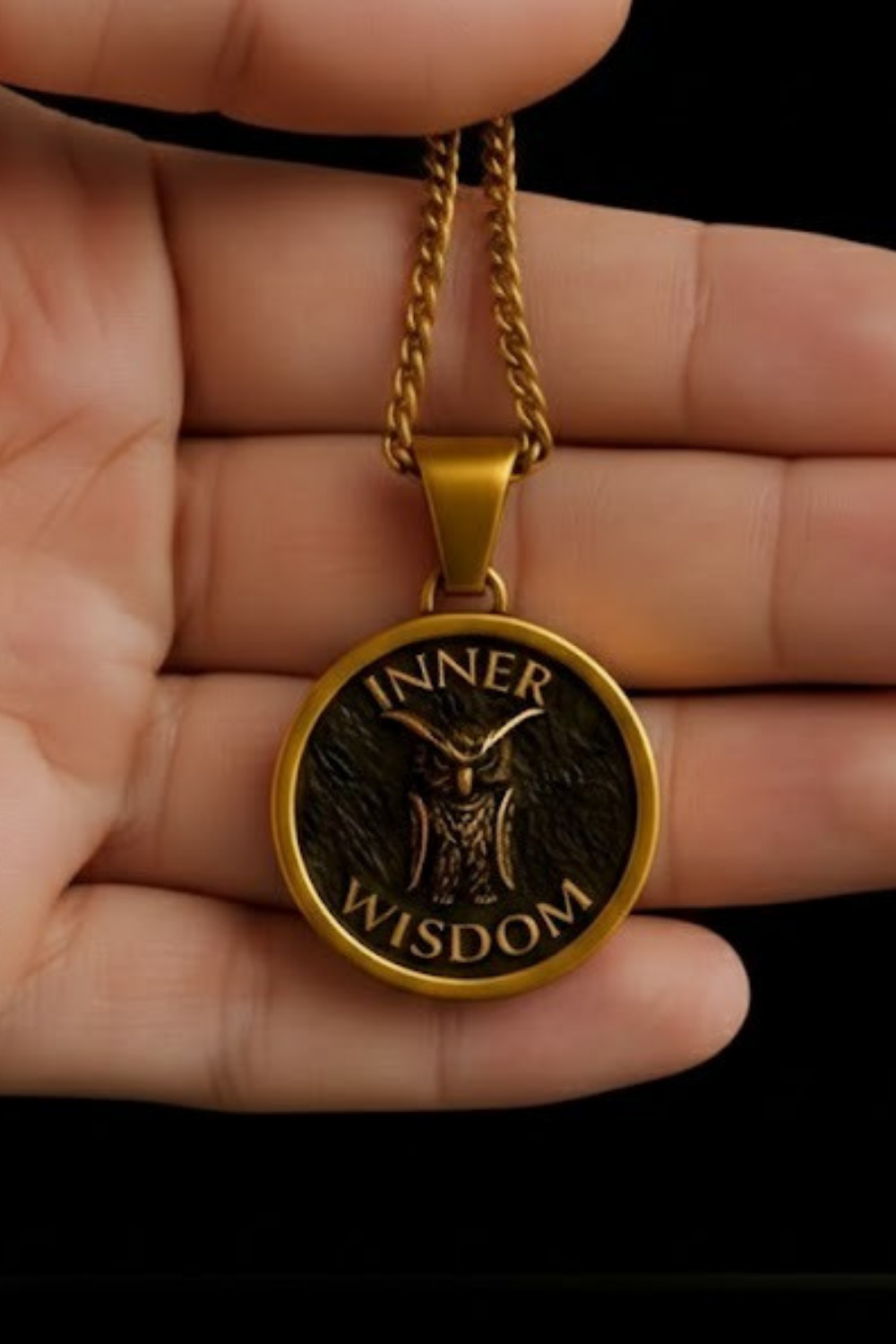 A gold-colored pendant necklace with a circular design featuring an owl and the text "INNER WISDOM" is being held in a person's hand.
