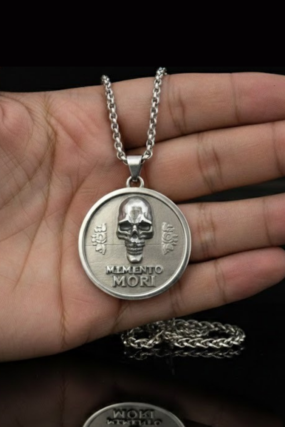 A silver pendant necklace with a circular medallion featuring a skull and the Latin phrase "Memento mori" (which means "Remembering the dead" in English).