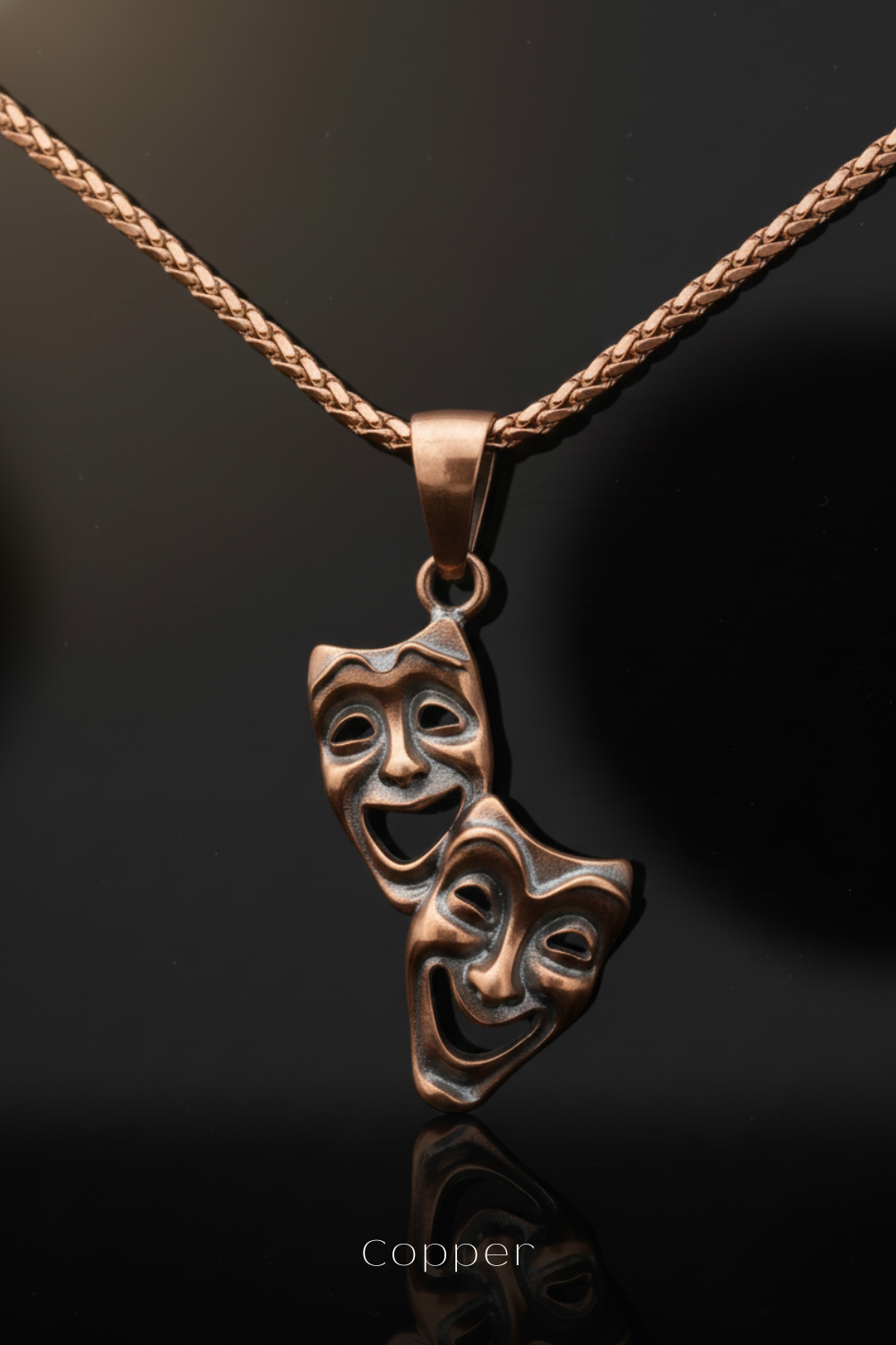 A copper-colored pendant necklace with two smiling theatrical masks hanging from it.