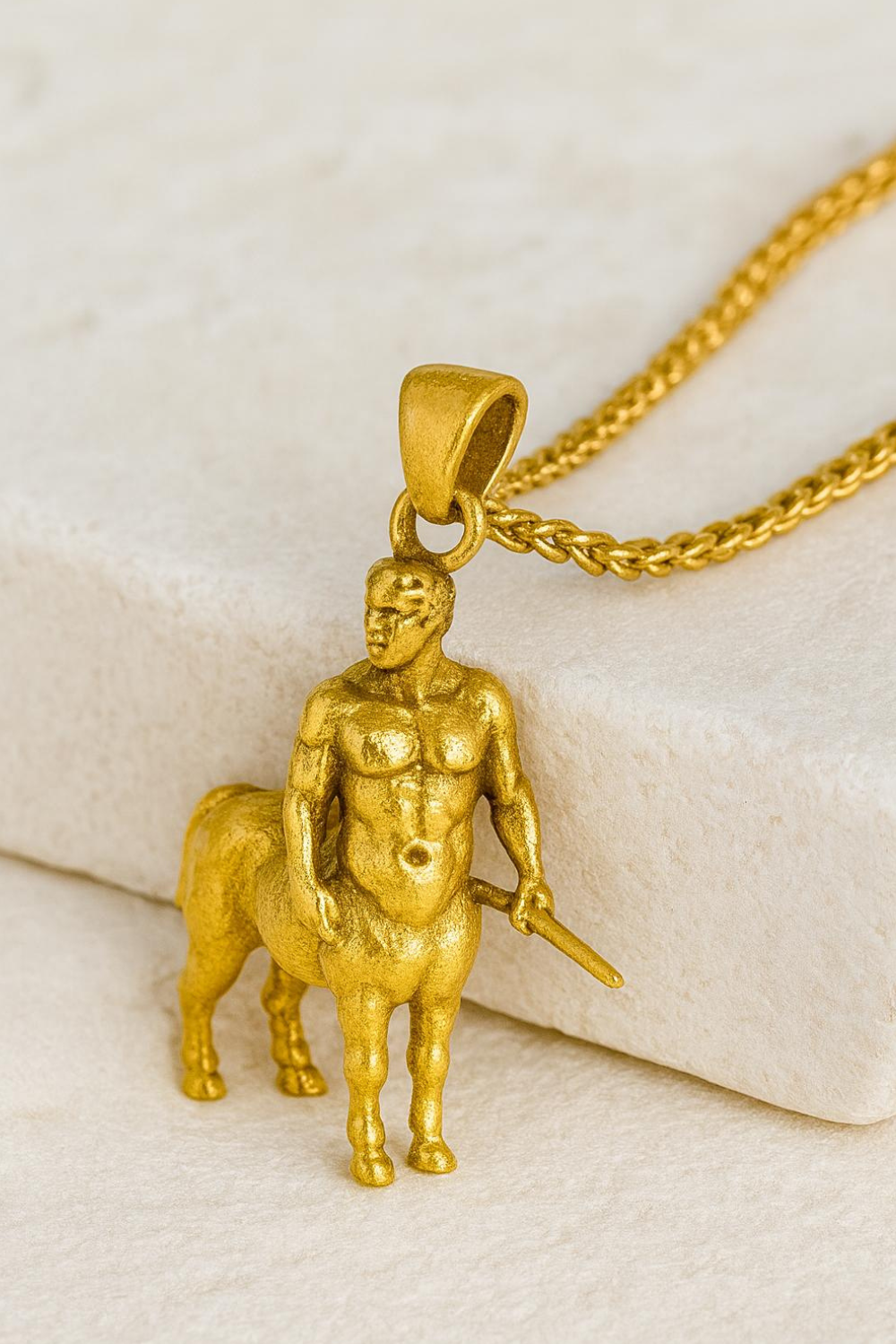 A gold-colored pendant in the shape of a muscular male figure, possibly a centaur, is displayed on a chain.
