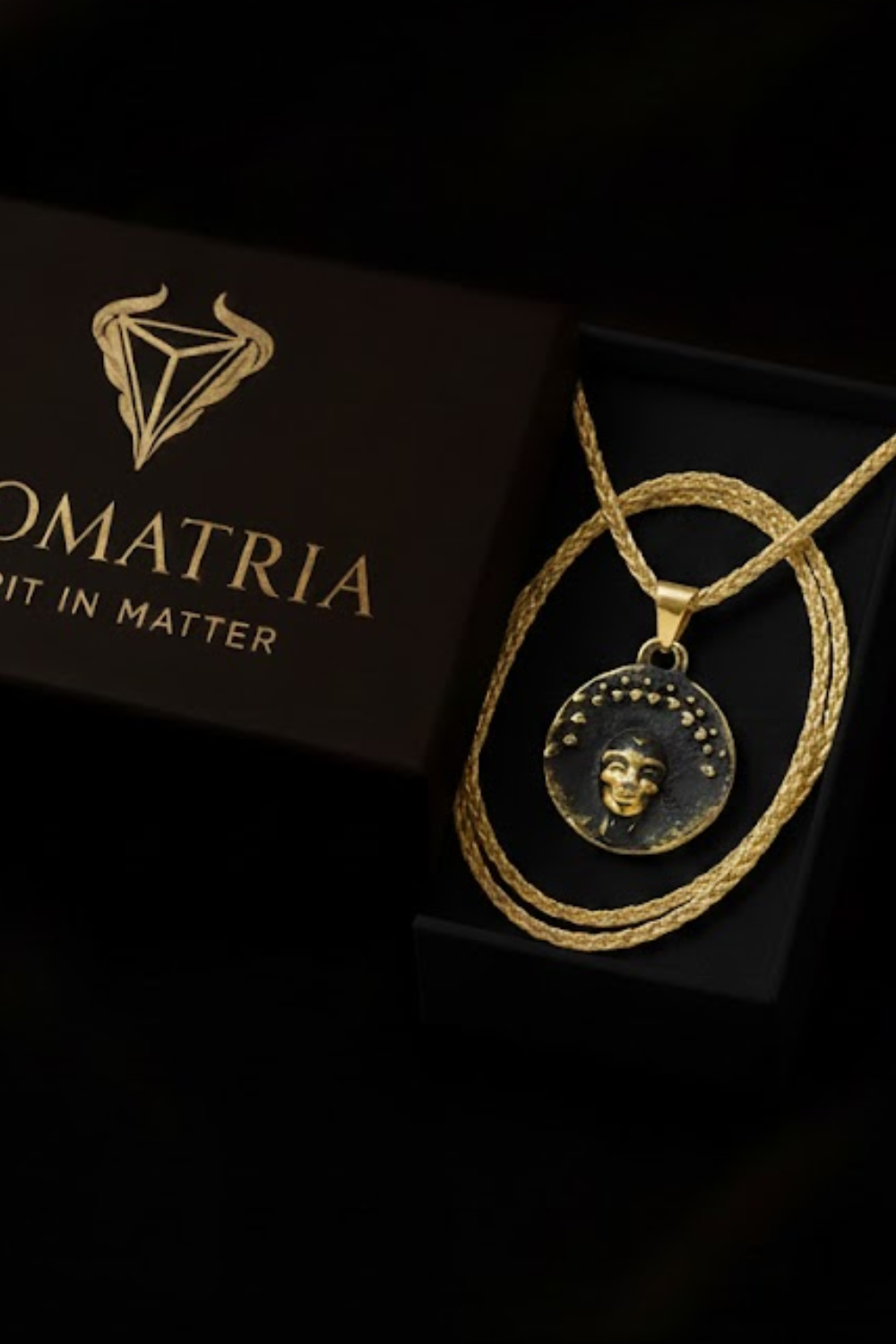 a pendant necklace with a gold-colored pendant, displayed in a black box with a gold-colored label that reads "OMATRIA".