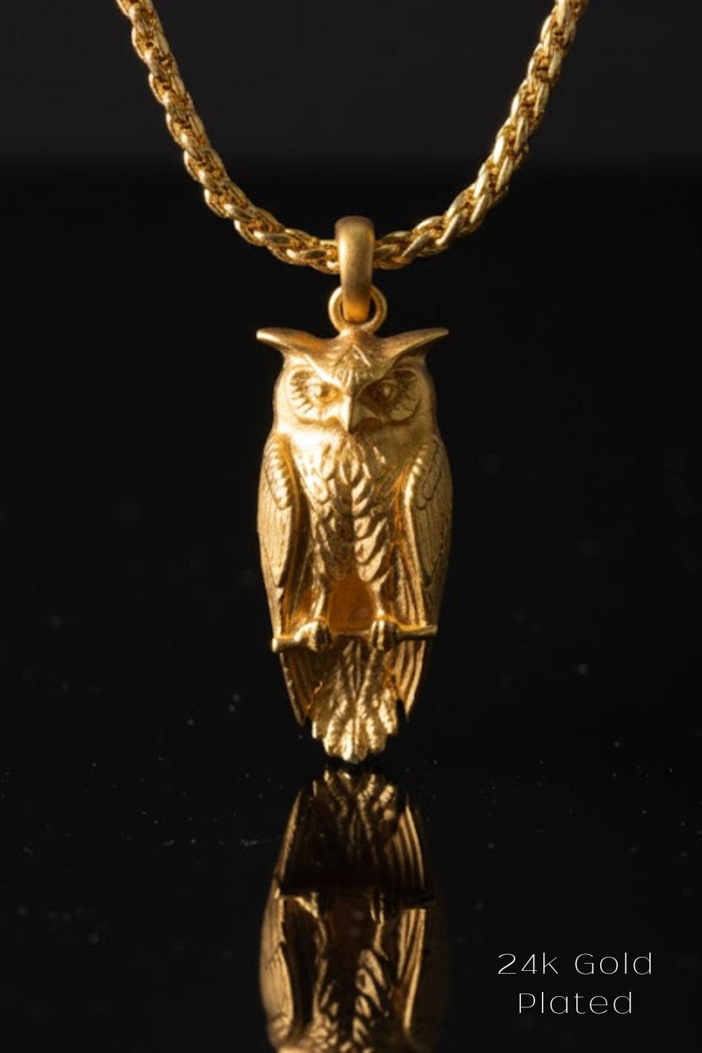A 24k gold plated pendant in the shape of an owl is displayed against a black background, with its reflection visible below it.
