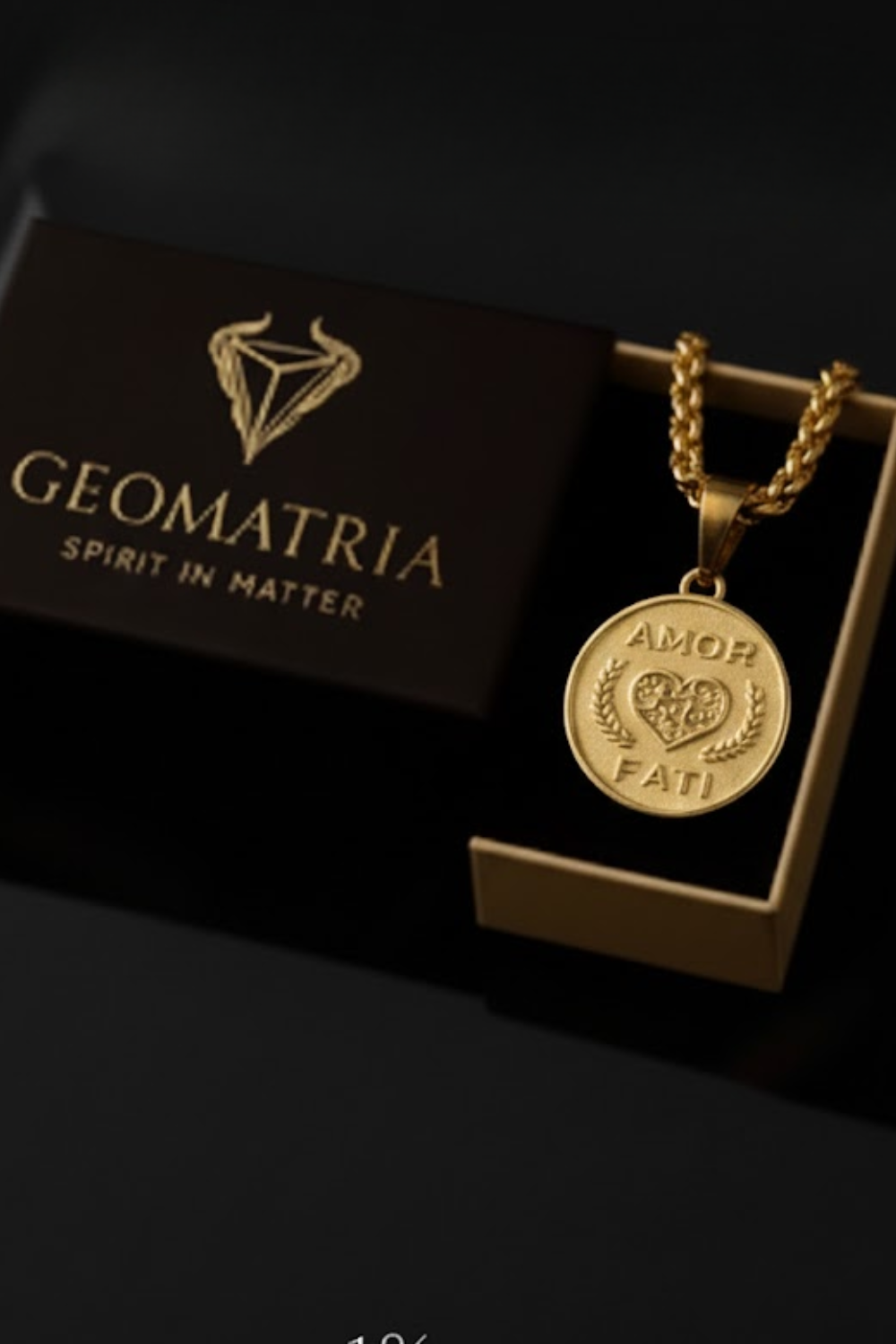 A gold-colored pendant necklace with a heart-shaped medallion attached, displayed in a black box with the Geomatria logo.
