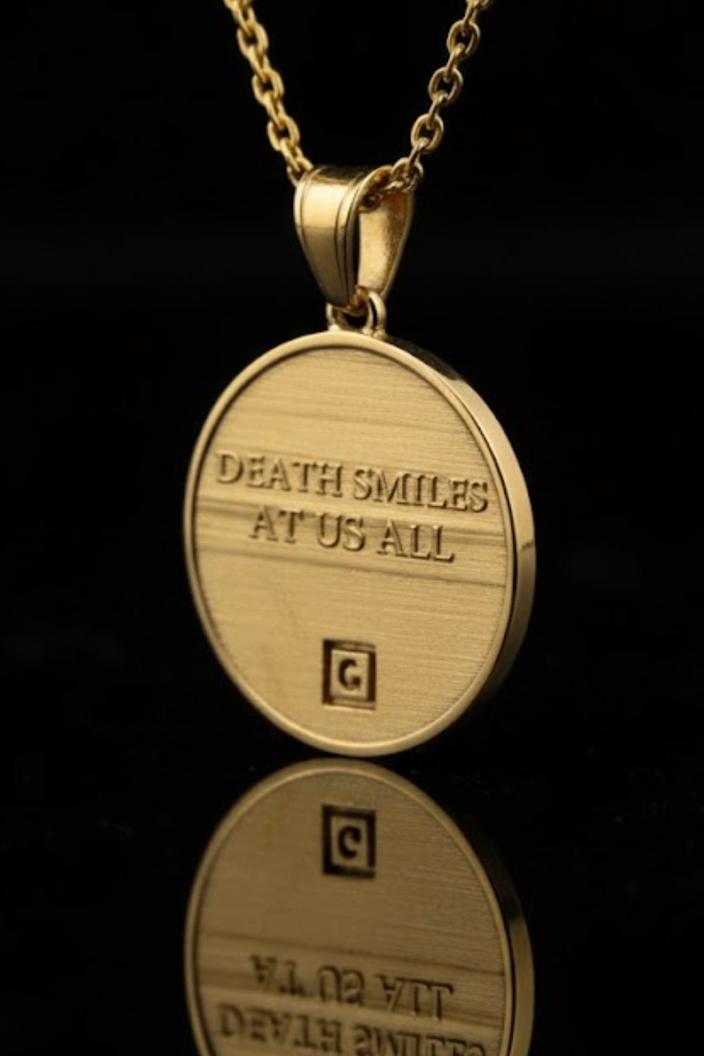 A gold-colored pendant necklace with a circular medallion-shaped design, featuring the text "DEATH SMILES AT US ALL" engraved on it.