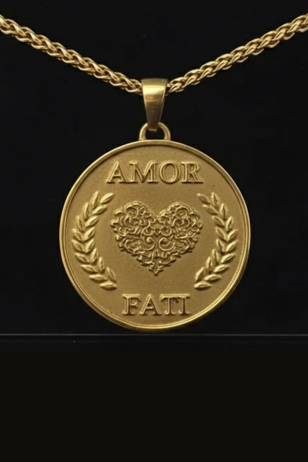 A gold-colored pendant necklace with a heart-shaped medallion featuring the words "Amor Fati" engraved on it.