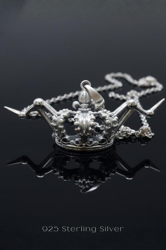 A silver-colored pendant necklace with a crown-shaped pendant is displayed against a dark background.