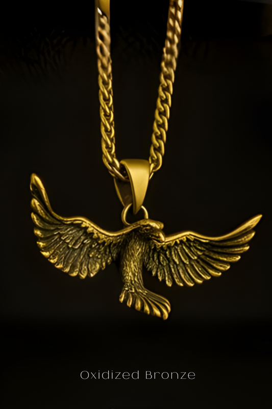A close-up view of a gold-colored pendant in the shape of a bird, with its wings spread wide, against a dark background.