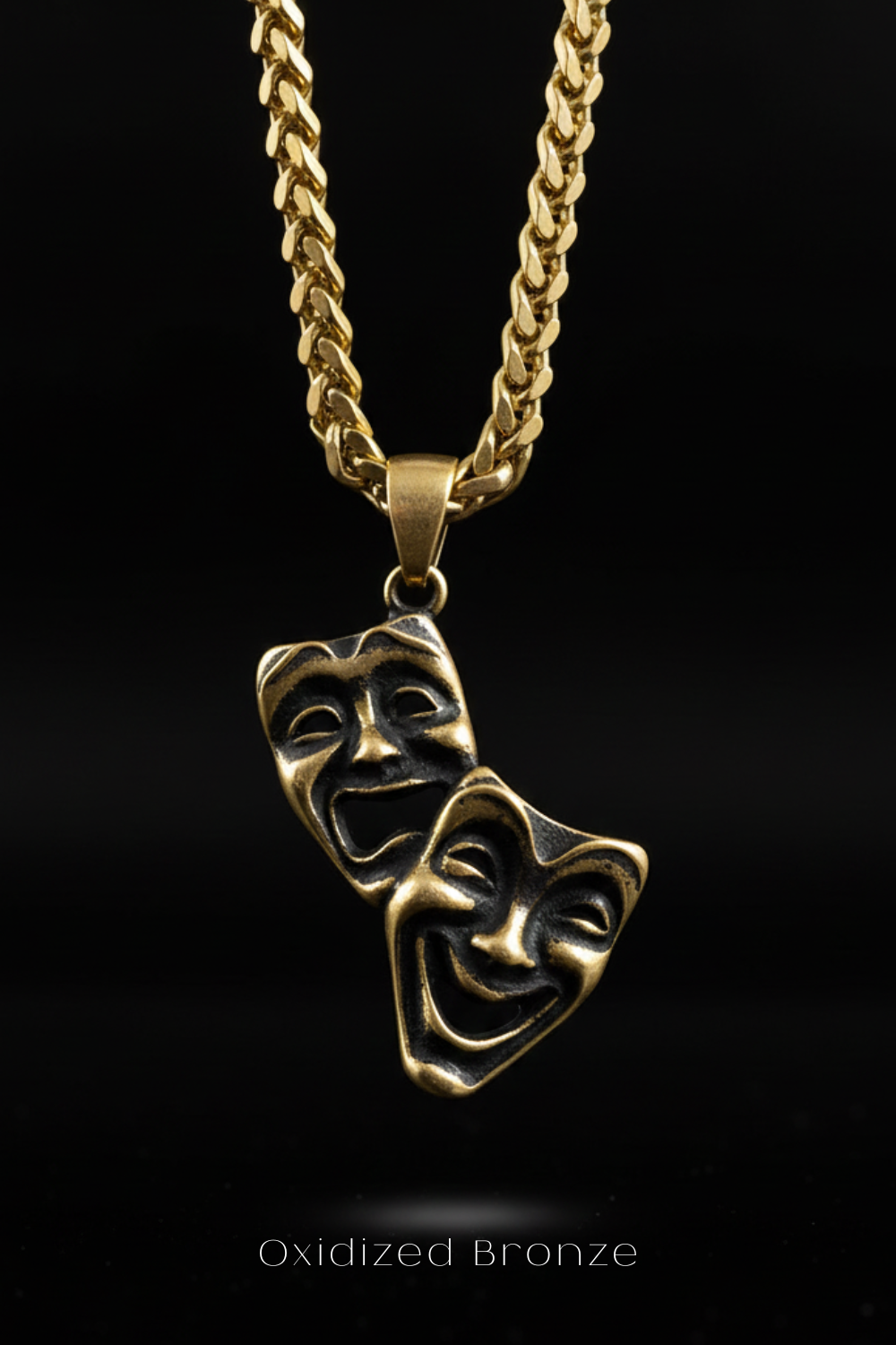 A gold-colored necklace with a pendant featuring two bronze-colored theatrical masks with exaggerated features, such as wide smiles and exaggerated eyes, against a black background.