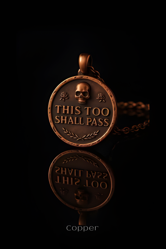 A copper-colored pendant with the text "THIS TOO SHALL PASS" engraved on it, along with a skull and rose design.