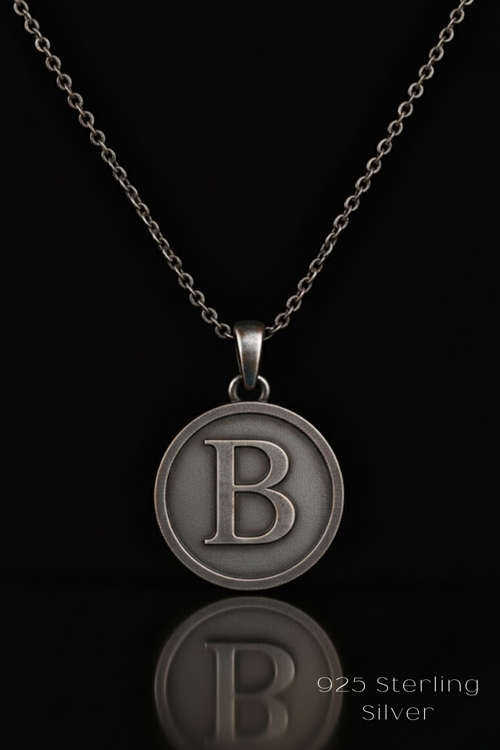 A silver-colored pendant necklace with a large letter "B" in the center, suspended from a chain against a black background.