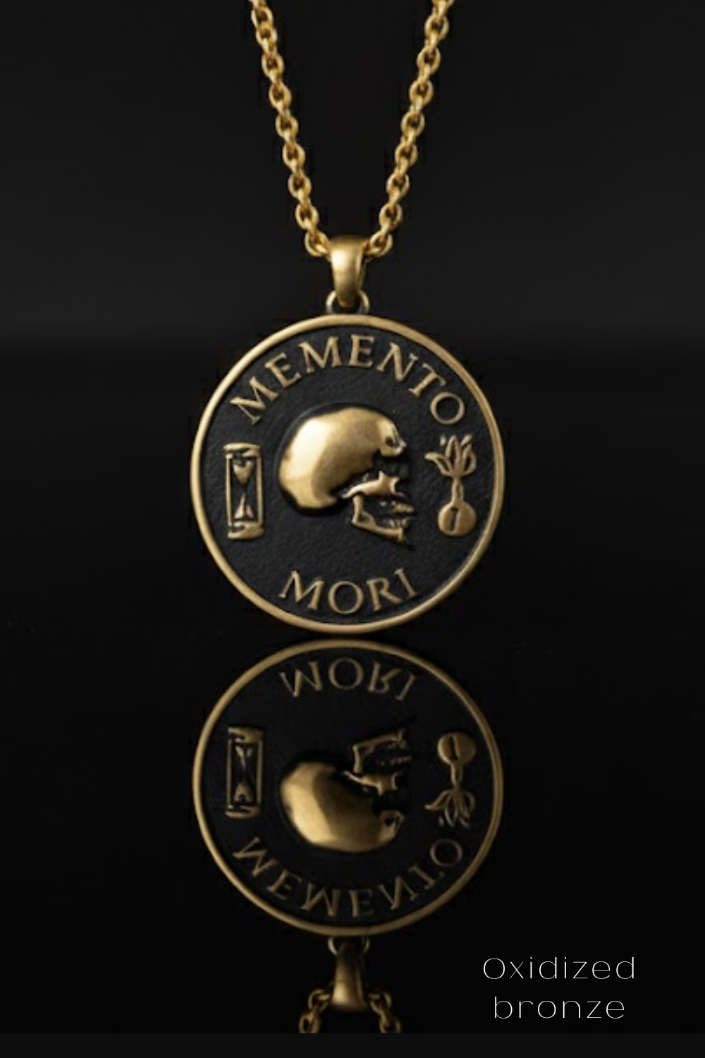 a gold-colored pendant necklace with a circular medallion that has a skull and the Latin phrase "Memento mori" inscribed on it. The pendant is displayed against a black background, and its reflection can be seen in the foreground.