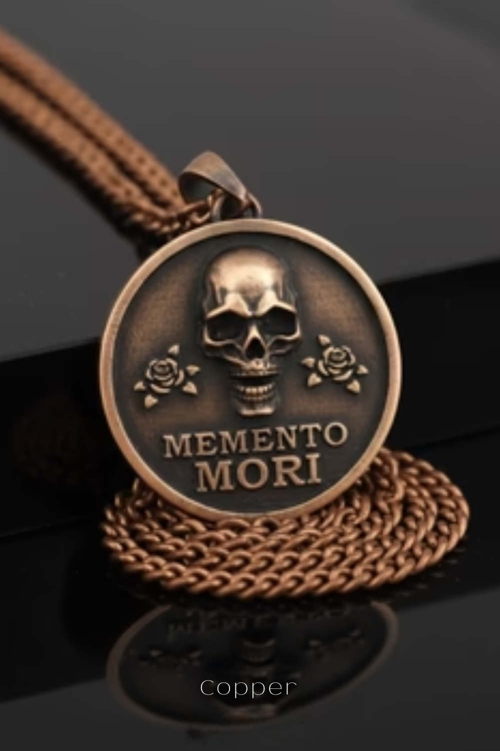 A bronze pendant with a skull design and the text "MEMENTO MORI" engraved on it, resting on a chain.
