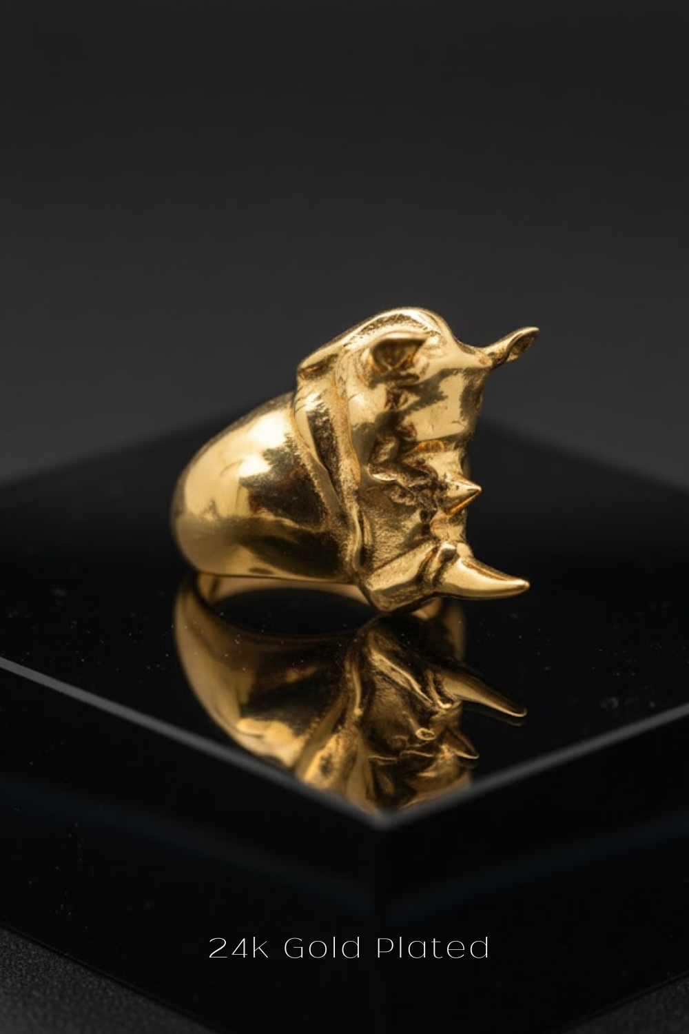A 24k gold plated ring with a unique, sculpted design sits on a black surface, reflecting the image.