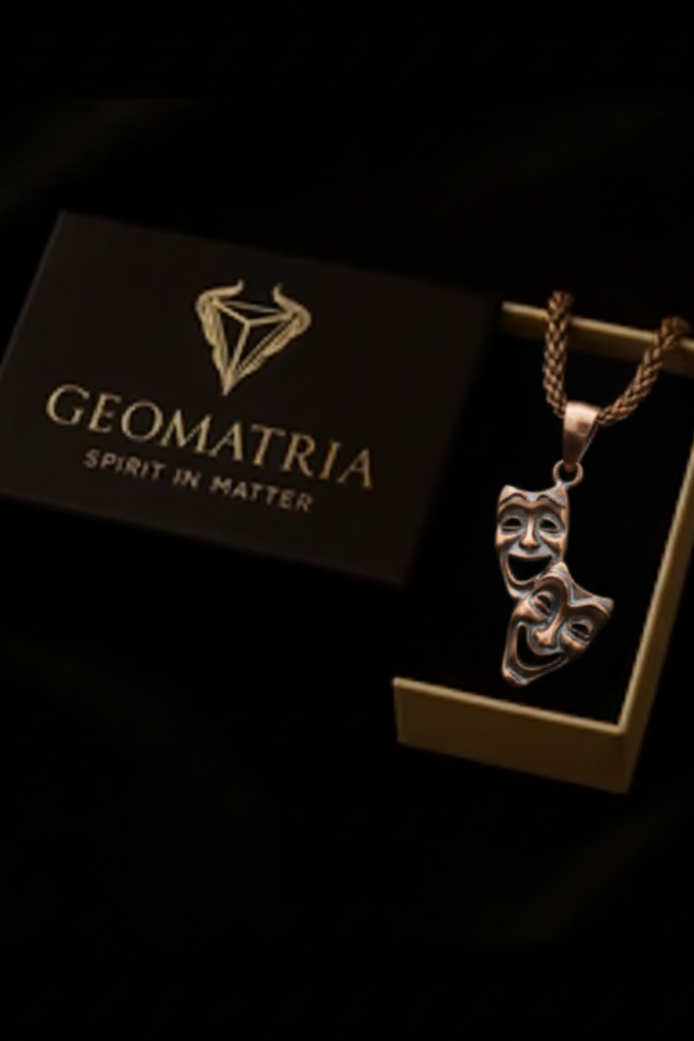 a necklace with a pendant in the shape of a theater mask, displayed in a box with the text "Geomatria Spirit in Matter".