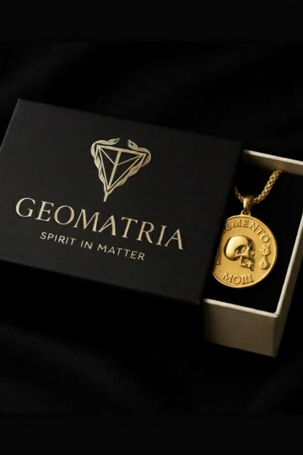 A gold-colored pendant necklace with a skull pendant is displayed in a black box with the text "Geomatria" and "SPIRIT IN MATTERS" printed on it.