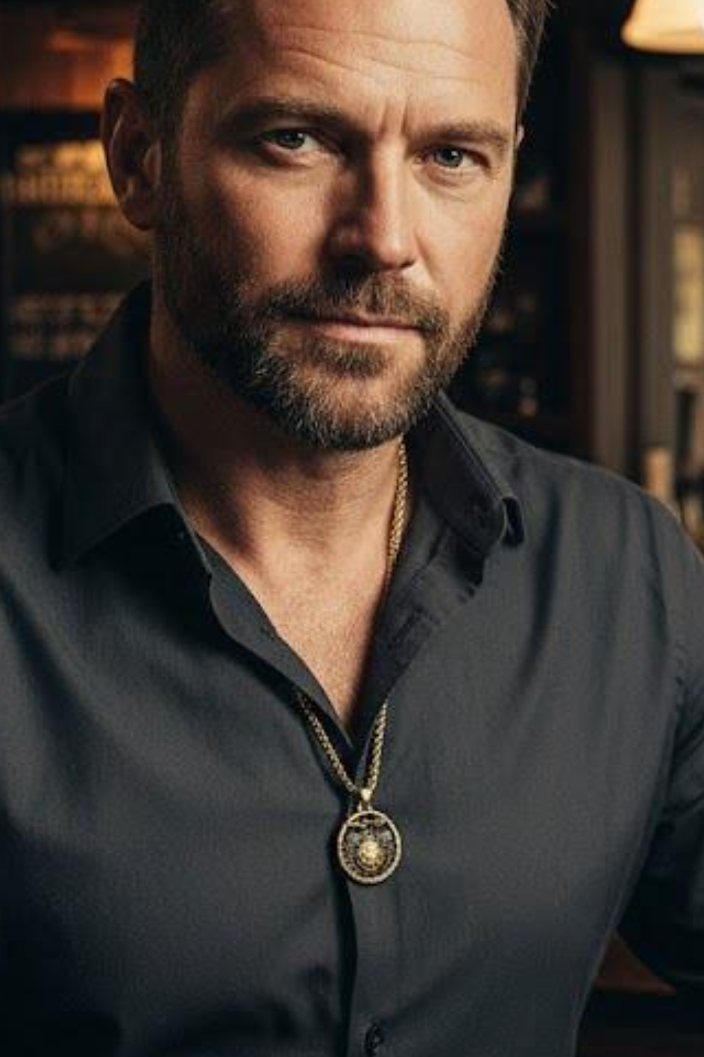 A man with a beard wearing a black shirt and a gold necklace is looking directly at the camera.