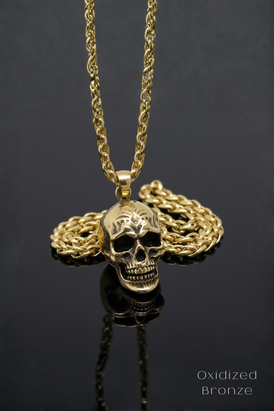 A gold-colored necklace with a pendant featuring a skull design, resting on a reflective surface.