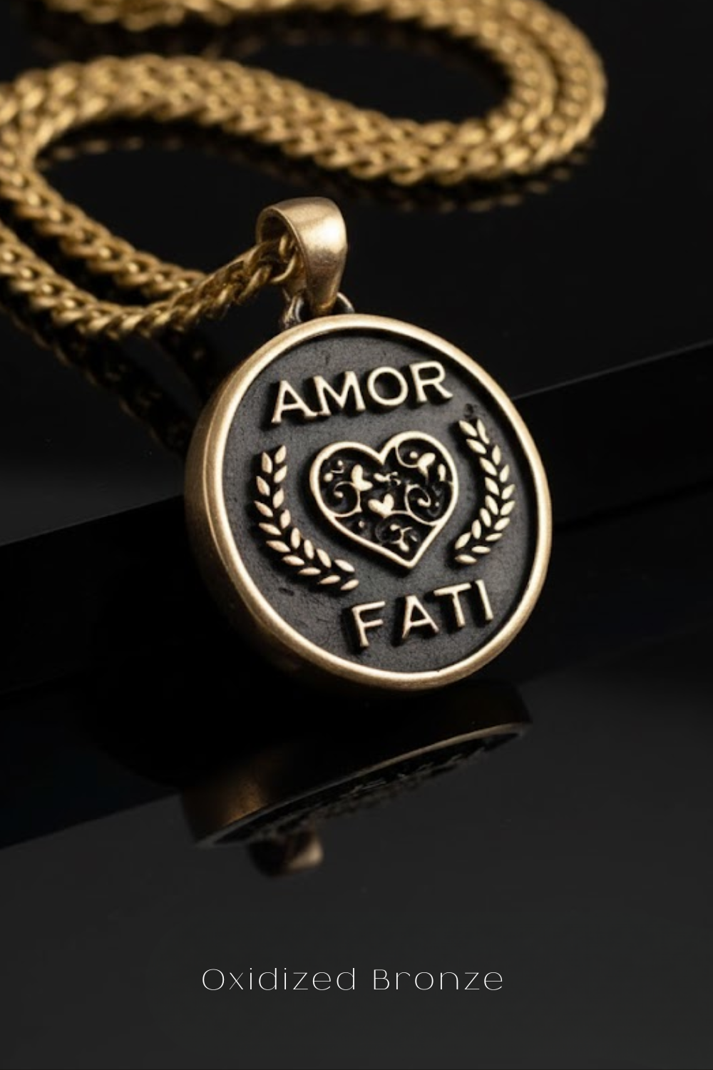 a close-up view of a pendant with a heart and laurel wreath design, attached to a gold chain. The pendant has the words "Amor Fati" engraved on it, which translates to "Love is a feast" in Italian. The pendant is placed on a reflective surface, creating a visually appealing contrast between the gold chain and the black background.
