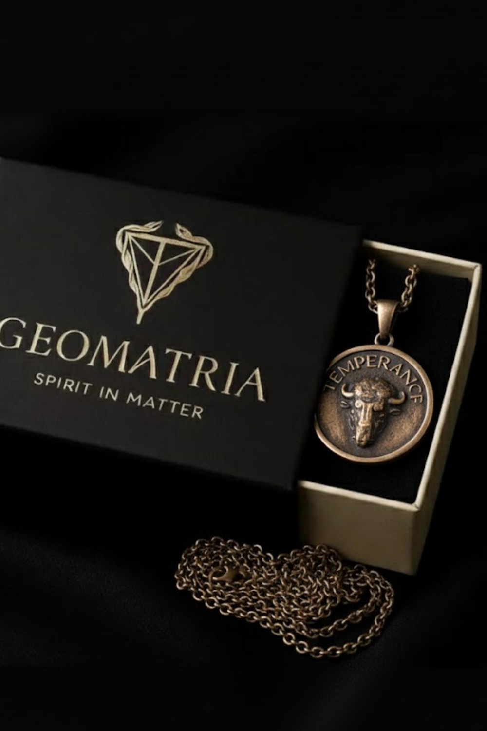 A black box with a gold geometric design and the text "GEOMATRIA SPIRIT IN MATER" on it, along with a gold-colored pendant featuring a bull's head design.