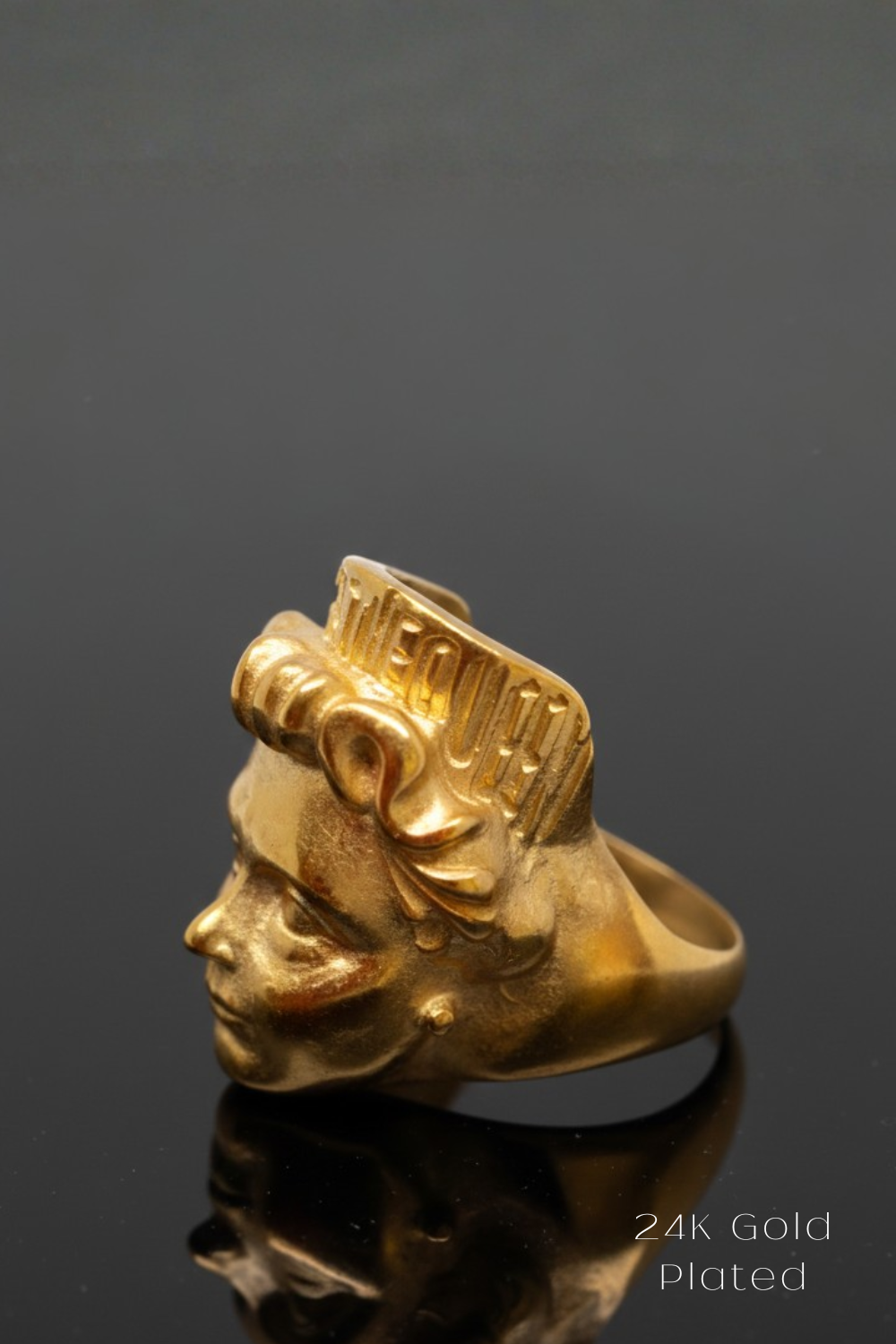 A gold-colored ring with a stylized profile of a person's face on it, placed on a reflective surface.