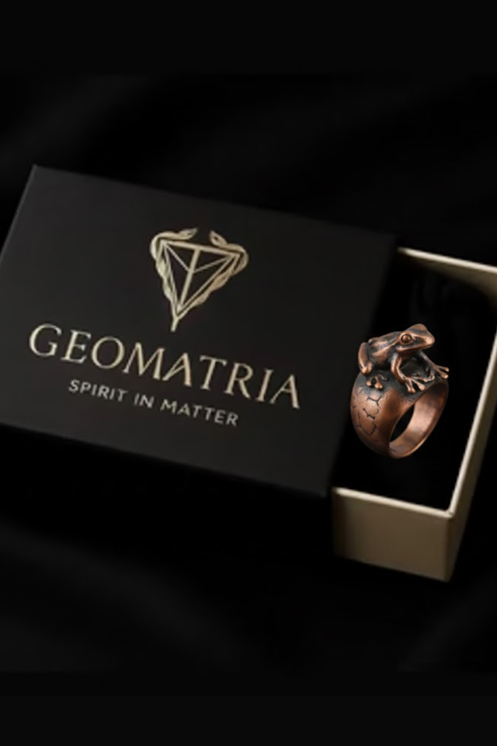 A black box with the word "Geomatria" printed on it, along with a copper-colored ring with a frog design inside the box.