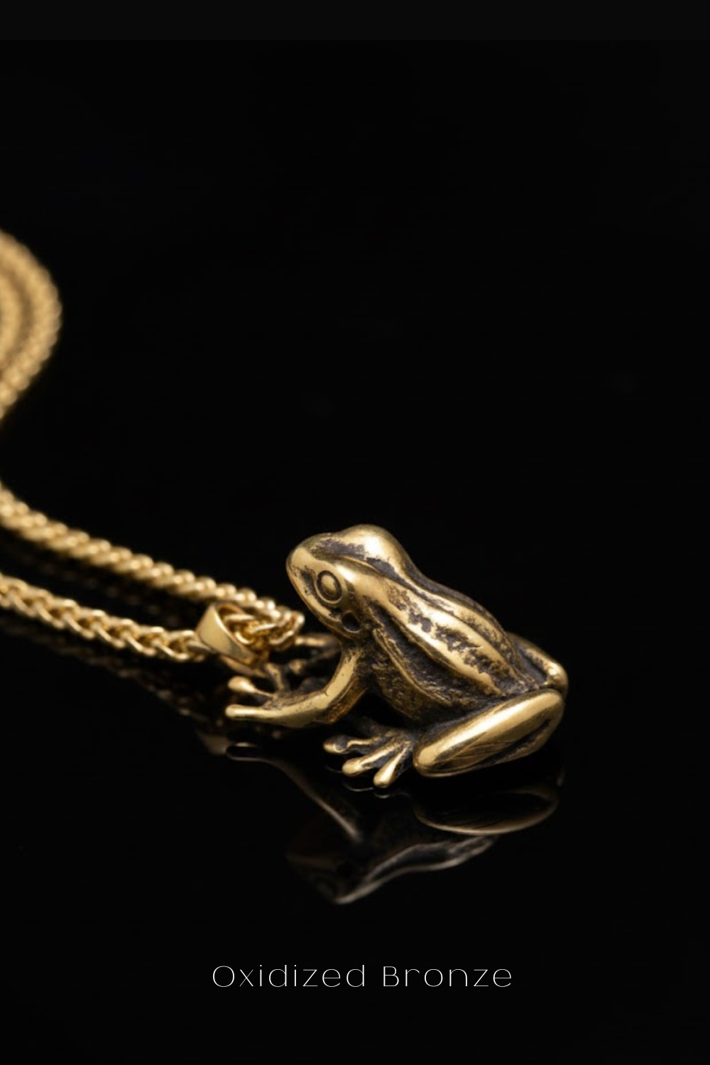 A close-up image of a gold-colored pendant in the shape of a frog, with a chain attached to it, against a black background.