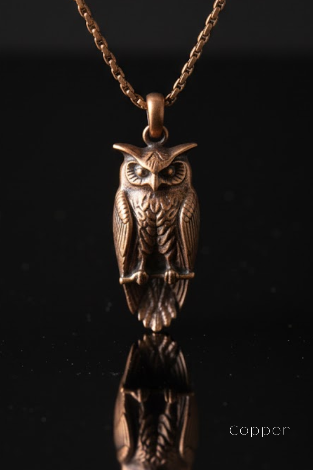 A copper-colored pendant in the shape of an owl, with a chain around it, is displayed against a black background.