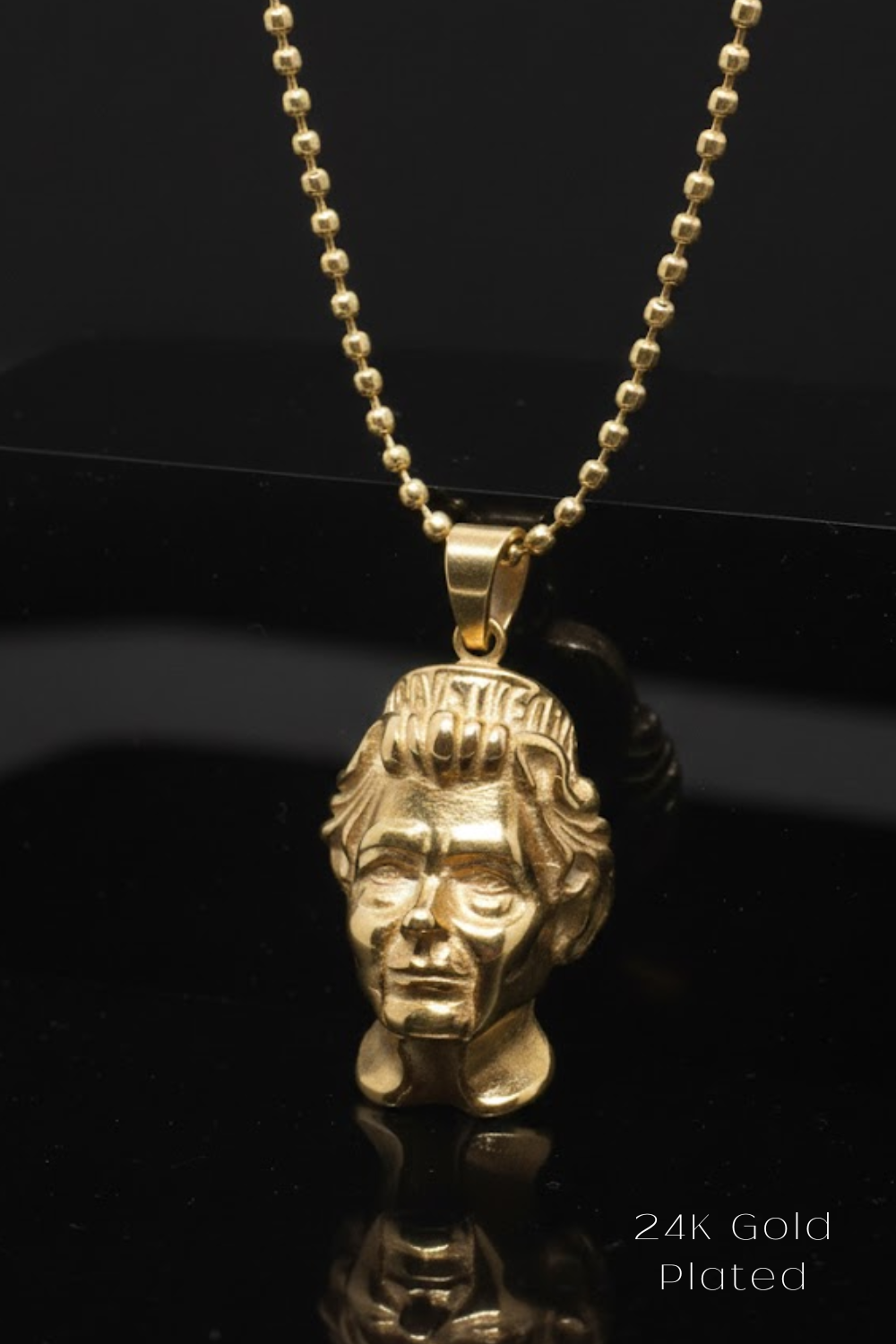 A gold-colored pendant in the shape of a human head, suspended from a chain against a black background.