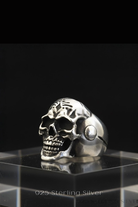 A silver skull ring is displayed on a clear glass stand against a black background.