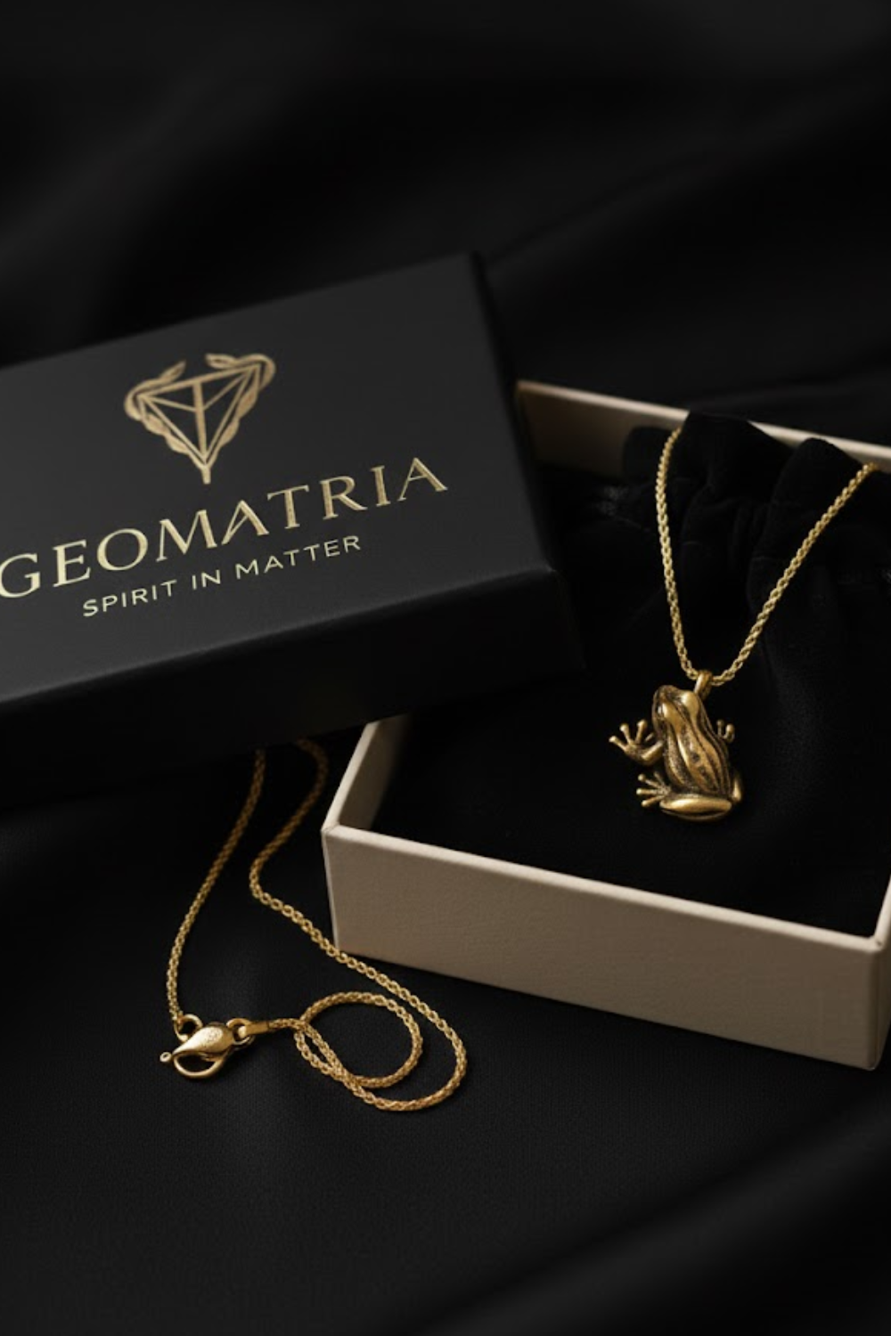 A gold-colored frog pendant necklace is displayed in a black jewelry box with a gold-colored geometric logo.