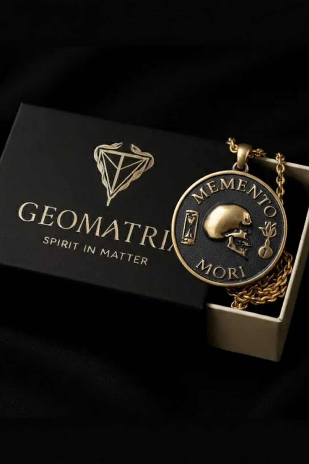 a gold-colored pendant with a black and gold design, including a skull and a plant, resting on a black box with the text "Geometria" and "Spirit in Matter" printed on it.