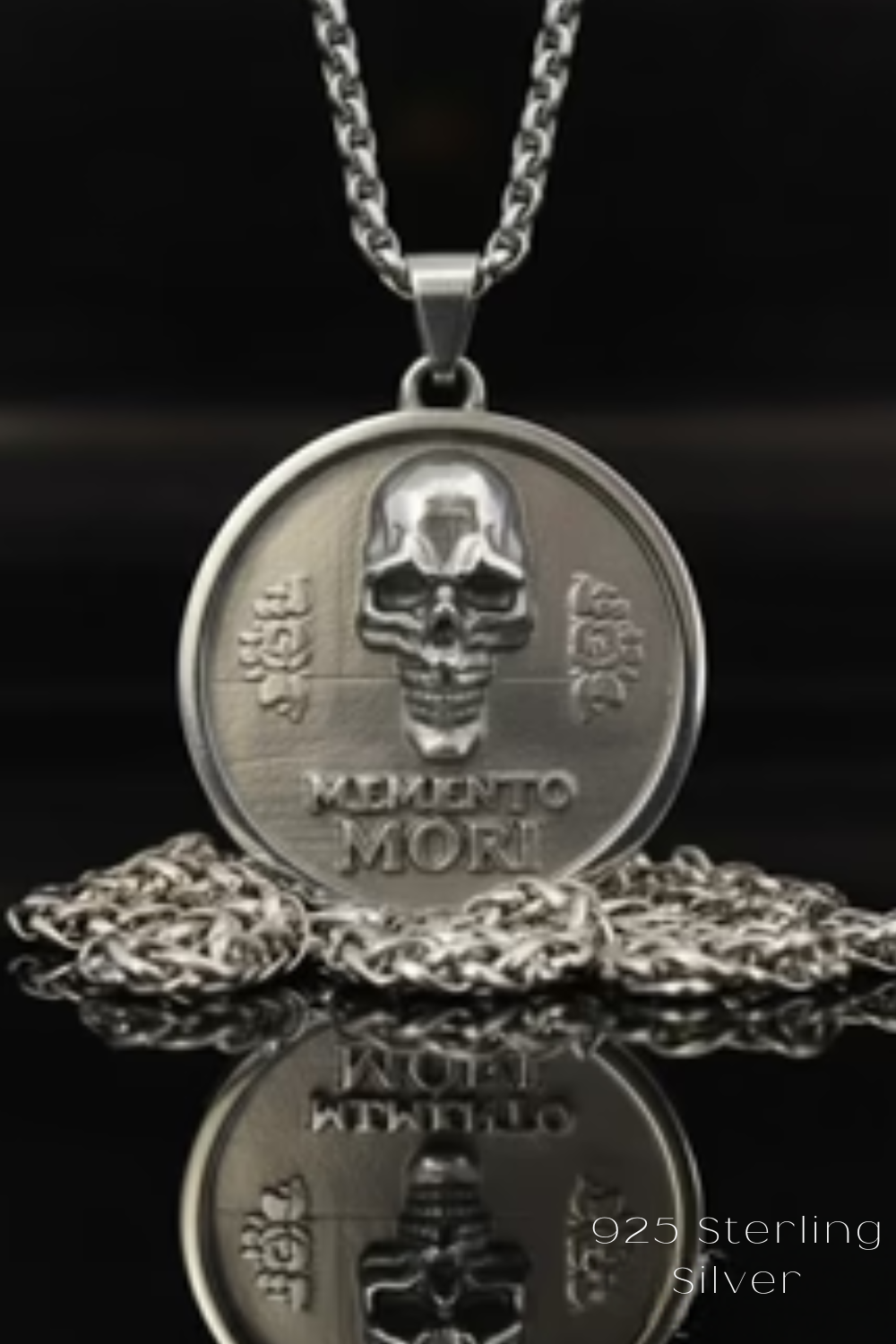 A silver pendant necklace with a skull design and the Latin phrase "Memento mori" engraved on it.