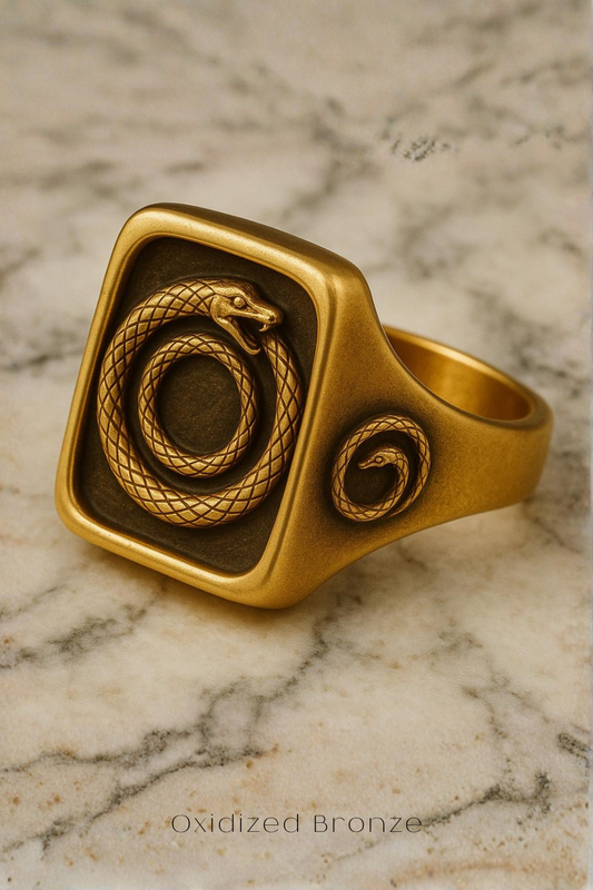 A gold ring with a square-shaped design featuring a snake and a smaller snake inside, resting on a marble surface.