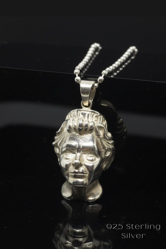 A silver pendant in the shape of a human head, with intricate details and a chain attached to it, is displayed against a black background.